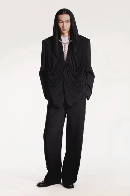 Asian male fashion model in black hooded draped jacket and wide-leg pants against white background in studio fashion shot.