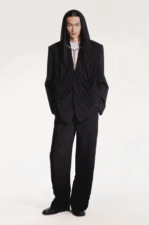 Asian male fashion model in black hooded draped jacket and wide-leg pants against white background in studio fashion shot.