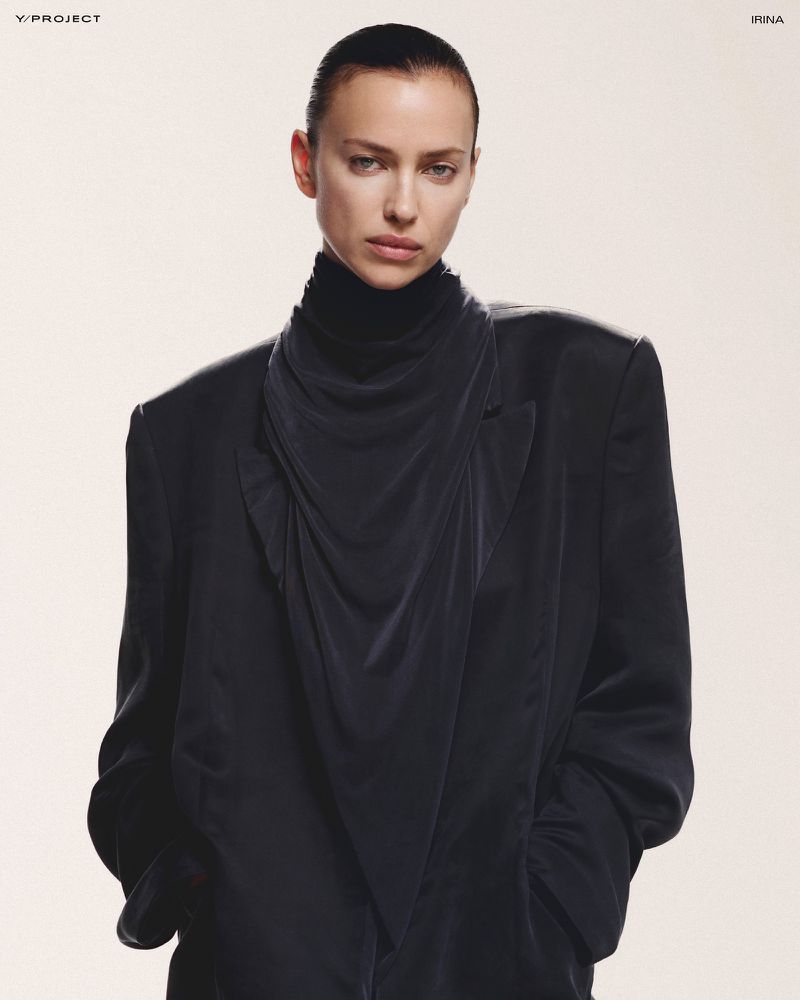 Elegant fashion portrait of Irina Shayk wearing a black turtleneck and draped blazer against a neutral backdrop in a studio environment.