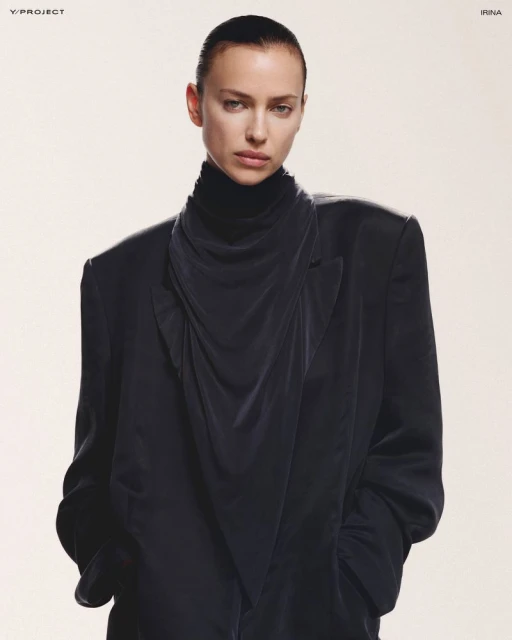 Elegant fashion portrait of Irina Shayk wearing a black turtleneck and draped blazer against a neutral backdrop in a studio environment.