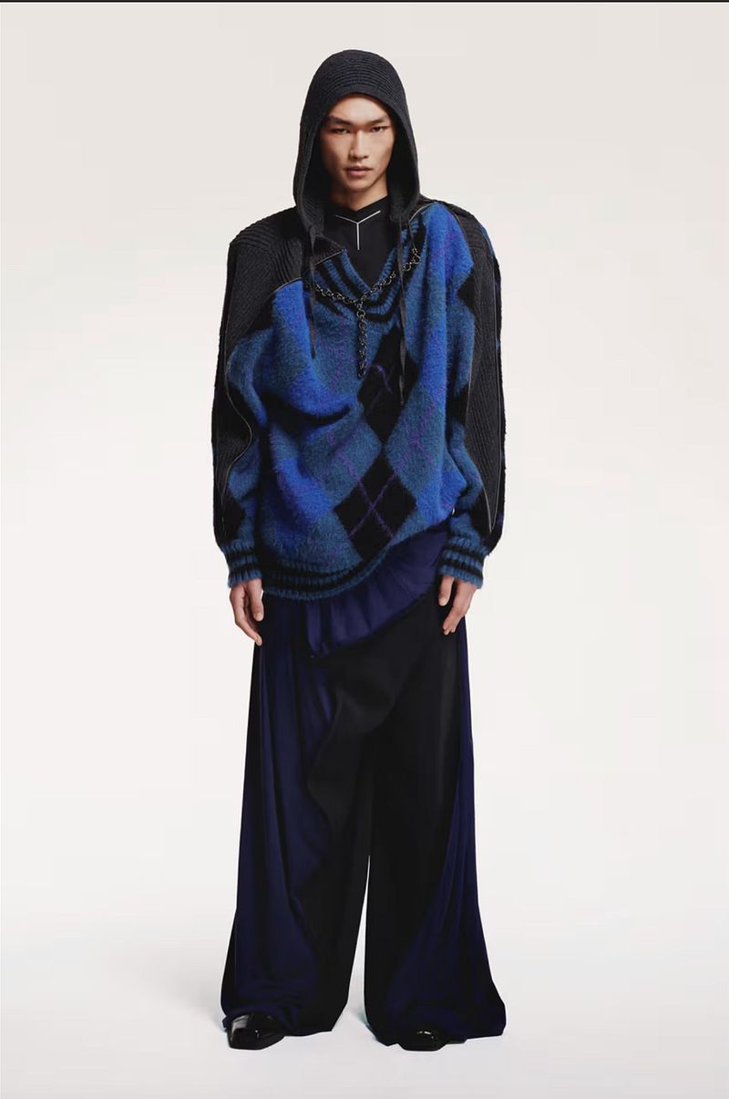 Full-length shot of a model in a studio wearing a blue argyle sweater over a black hooded jacket and draped wide leg pants.