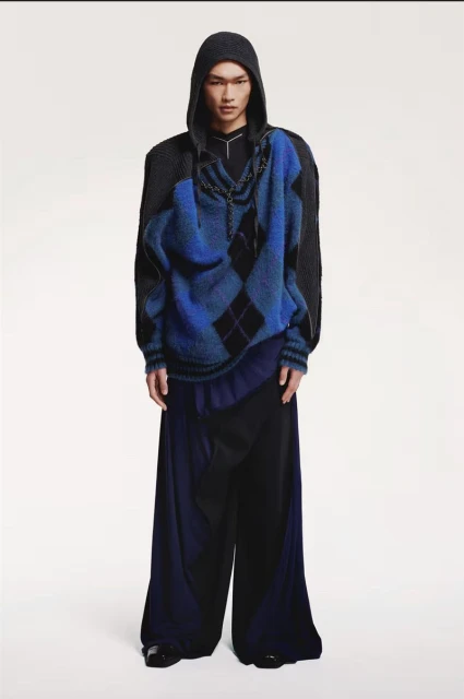 Full-length shot of a model in a studio wearing a blue argyle sweater over a black hooded jacket and draped wide leg pants.