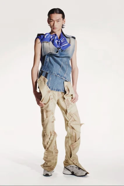 An Asian man stands in a sleeveless denim top and draped khaki pants in a full shot against a bright white studio background.