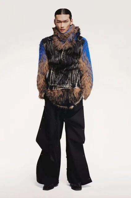 An adult male model poses in a studio wearing a black and tan fur-trimmed vest, an azure and tan fur sweater, and black wide-leg pants.