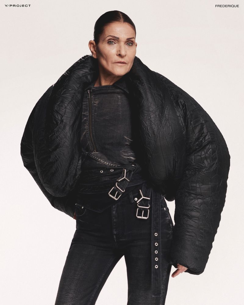 Adult woman in a black leather jacket and jeans poses indoors against a plain white background in a fashion studio shot.