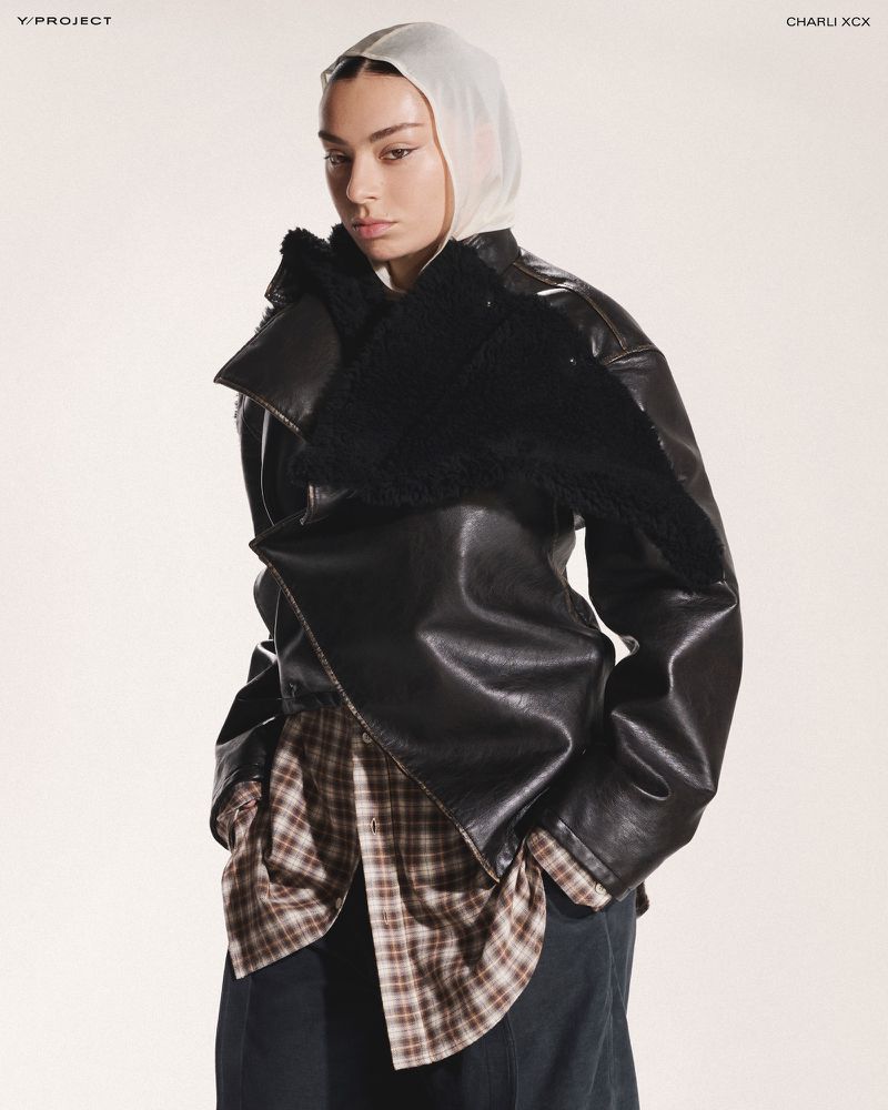 Fashion-forward adult female poses indoors wearing a black leather jacket with a shearling collar, plaid shirt, wide trousers, and a sheer headscarf.