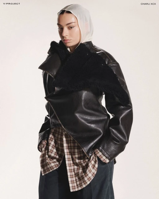 Fashion-forward adult female poses indoors wearing a black leather jacket with a shearling collar, plaid shirt, wide trousers, and a sheer headscarf.