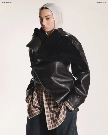 Fashion-forward adult female poses indoors wearing a black leather jacket with a shearling collar, plaid shirt, wide trousers, and a sheer headscarf.