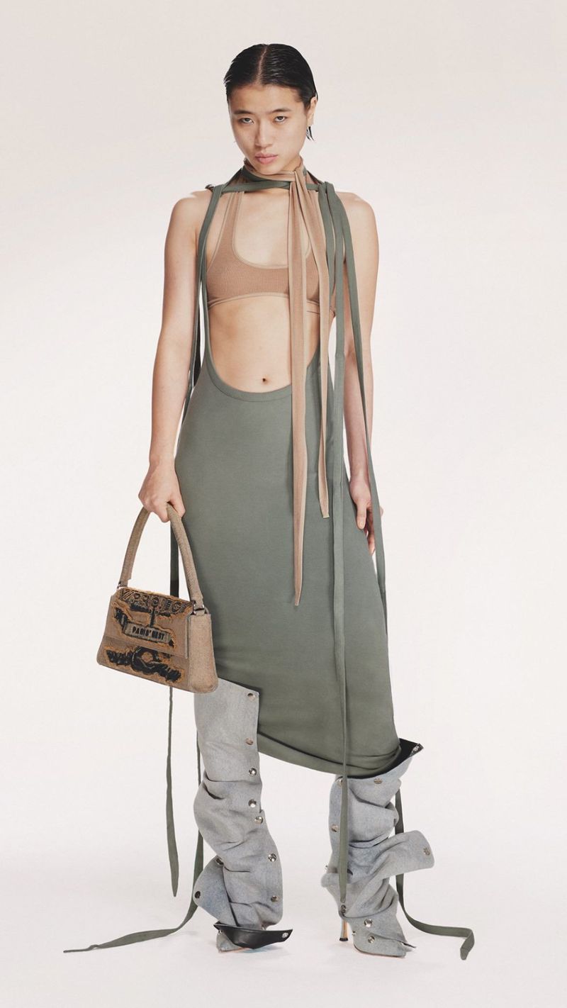 Fashion shot: an Asian woman wearing a green dress, tan top, decorative legwear and carrying a tan purse stands against a white background.