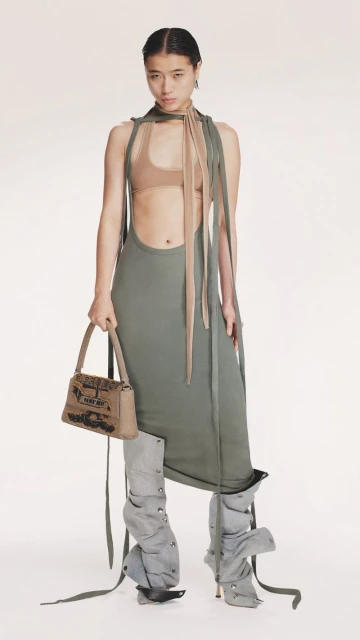 Fashion shot: an Asian woman wearing a green dress, tan top, decorative legwear and carrying a tan purse stands against a white background.