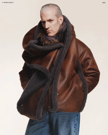 Editorial fashion shot of a grey-haired adult male model wearing a brown leather jacket with a grey fur collar and blue jeans in a studio.