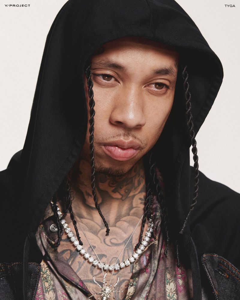 Portrait of Tyga wearing a black hoodie and diamond necklace against a neutral background, showcasing a mix of urban style and luxury.