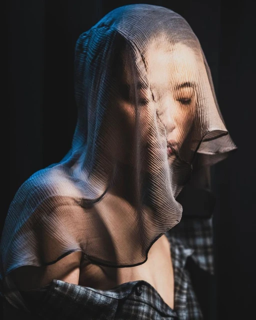 Close-up portrait of a woman with a sheer fabric draped over her head, wearing a plaid shirt against a dark background.