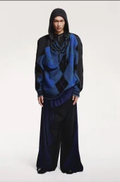 Model wearing a blue argyle sweater with a dark hooded top and wide trousers against a white background