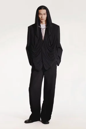 Model wearing a black hooded blazer with draped fabric and trousers against a clean white studio background.