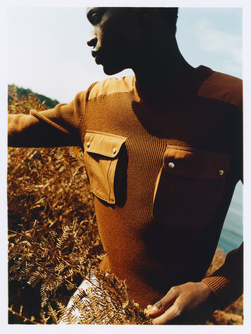 Man in tan sweater on an outdoor hillside. He is surrounded by dry foliage and a glimpse of ocean under a sunny sky.