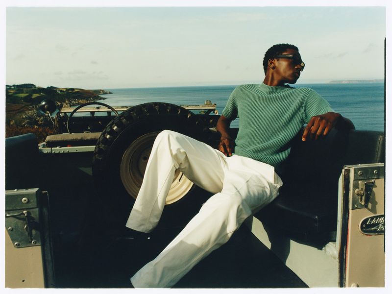 Fashion shot: an African-American man in sunglasses and a light-green shirt sits on the back of a Land Rover against a coastal backdrop.