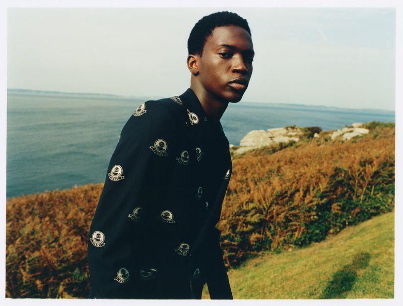 Outdoor fashion shot of a young Black man in a patterned shirt, standing on a grassy hill overlooking the ocean on a clear day.