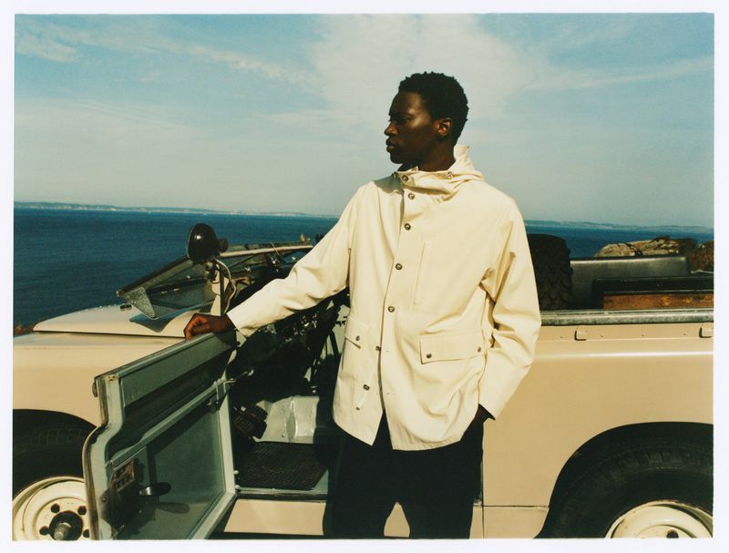A man in a cream-colored jacket stands next to a beige Land Rover overlooking the ocean with blue sky, creating a calm, outdoor scene.