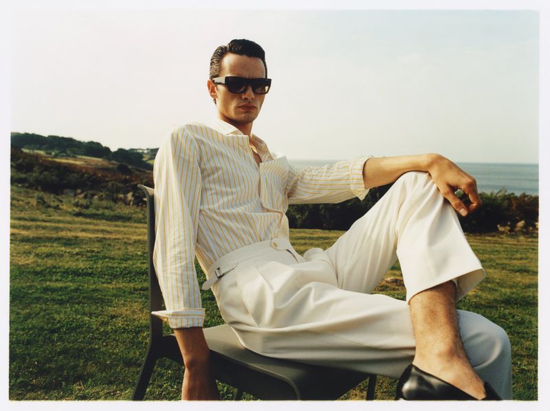 Man wearing sunglasses, striped shirt, and white pants poses while sitting on a chair outdoors in front of a grassy field and water.