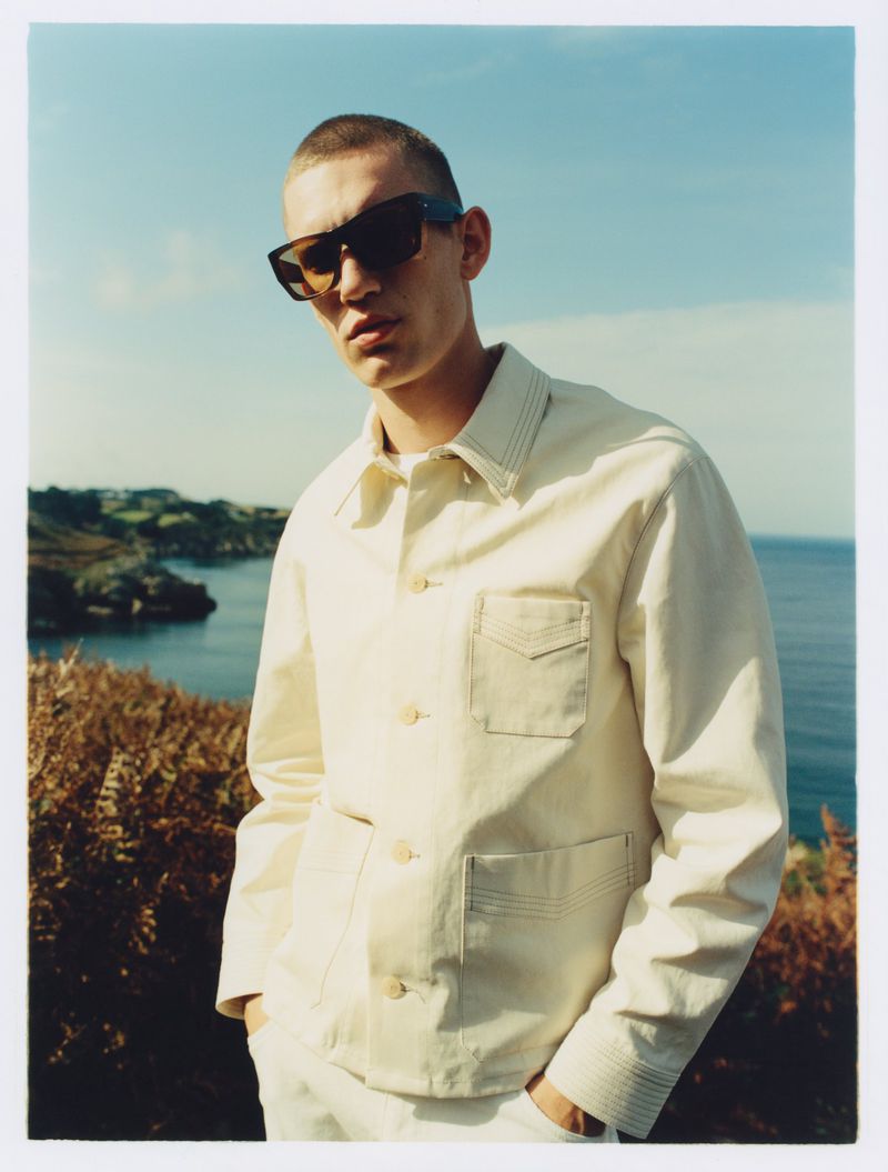 Man in sunglasses and beige jacket stands outdoors with coastline in background, showcasing fashion and travel style under natural light.