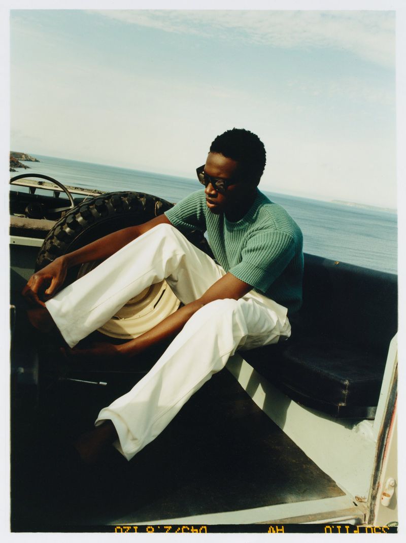 Man in sunglasses and casual wear sits in a vehicle with a spare tire, ocean and cloudy sky visible in the background.