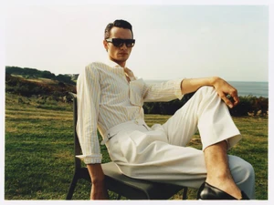 Man wearing sunglasses, striped shirt, and white trousers seated on a chair outdoors near the coast for Fursac S/S 24