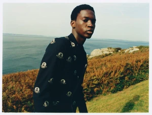 A Black man posing outdoors near a coastal landscape wearing a dark patterned shirt for Fursac S/S 24