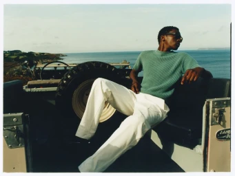 A man wearing a green knit shirt and white trousers seated in the back of an open vehicle overlooking the ocean.