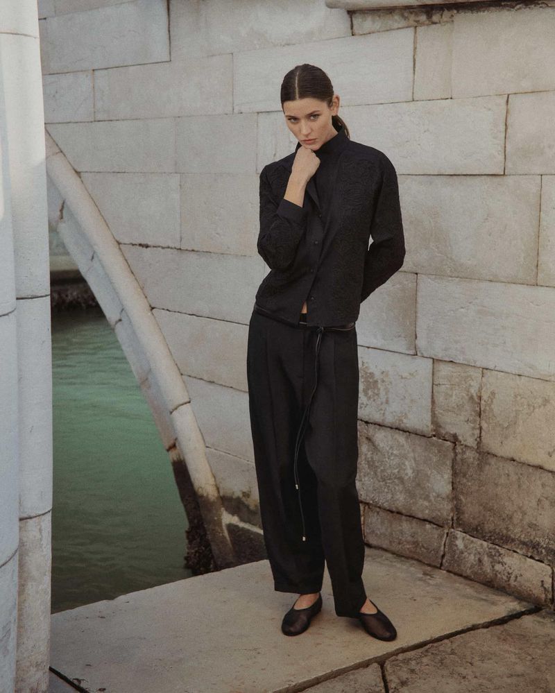 Elegant woman in a black outfit posing on a stone bridge with a waterway in the background; fashion editorial photography in Venice.