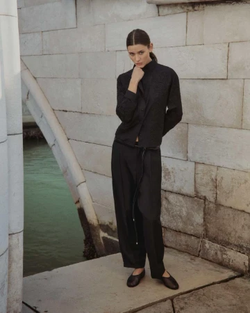 Elegant woman in a black outfit posing on a stone bridge with a waterway in the background; fashion editorial photography in Venice.