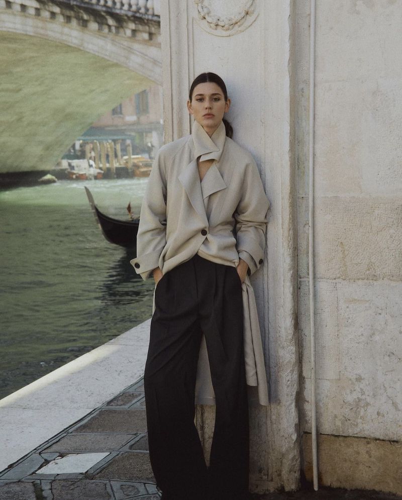 Brunette woman in a beige coat poses against an ornate wall with a bridge and canal visible in the background in Venice, Italy.