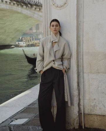 Brunette woman in a beige coat poses against an ornate wall with a bridge and canal visible in the background in Venice, Italy.