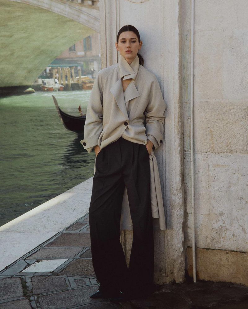 A young woman in a stylish tan coat and dark pants poses against a light-colored wall by a canal in Venice, Italy, under a bridge.
