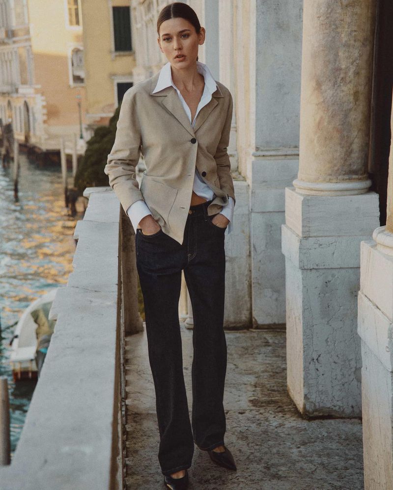 An adult woman in a blazer and jeans stands in front of a canal and buildings in Venice, Italy on a bright day.