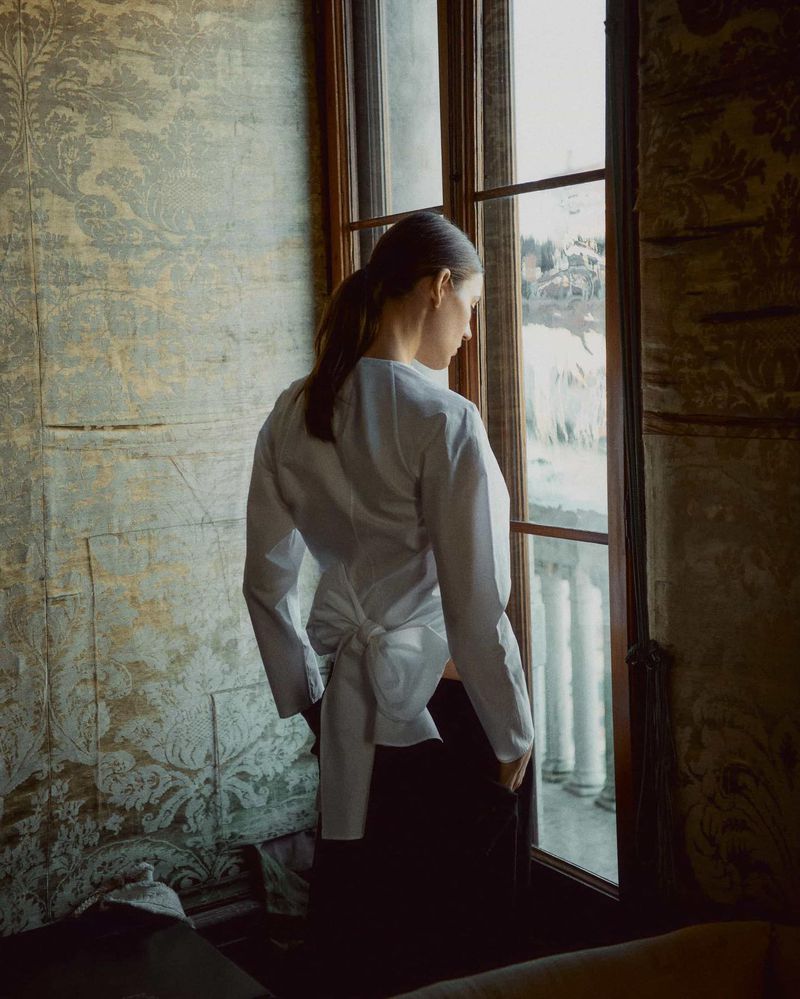 Indoor portrait of a woman in a white shirt looking out a window with light filtering in. Ornate, vintage wallpaper.