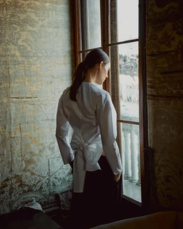 Indoor portrait of a woman in a white shirt looking out a window with light filtering in. Ornate, vintage wallpaper.