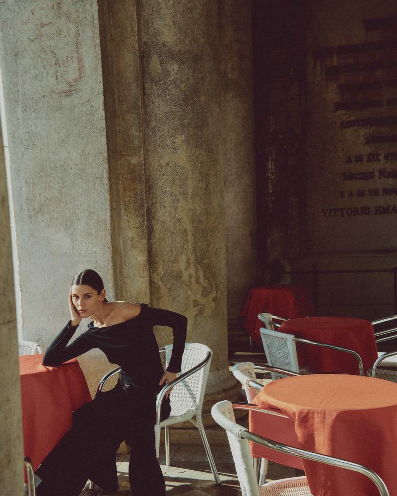 A woman in a black outfit sits near tables with red tablecloths under an arched stone structure. The shot is medium and the lighting is soft.