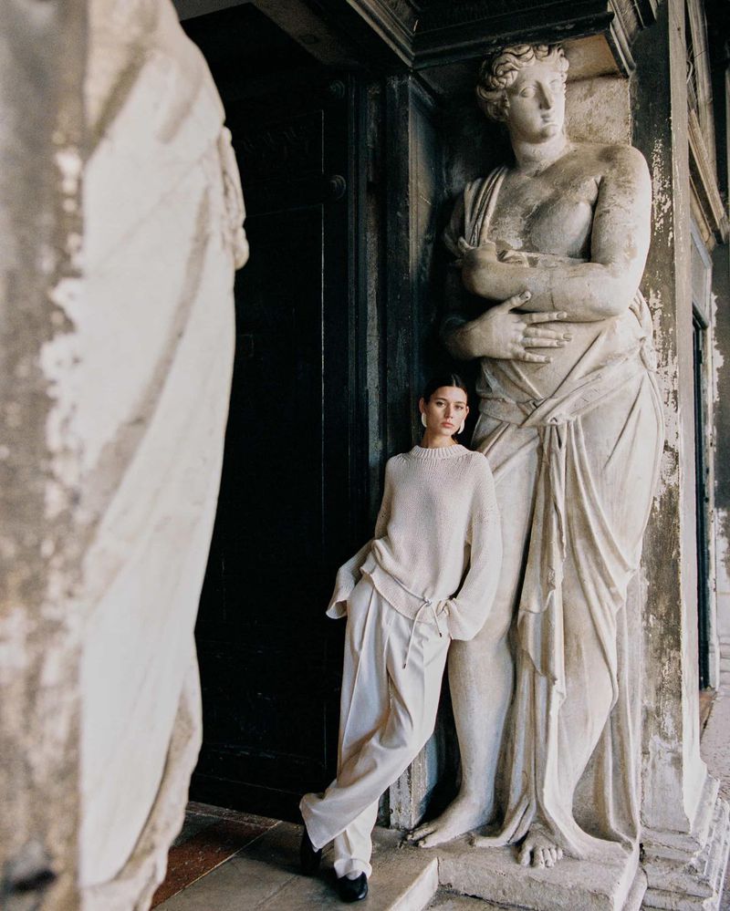 Woman in off-white sweater and pants poses next to a statue. Indoor shot with an architectural backdrop, soft light, and neutral colors.