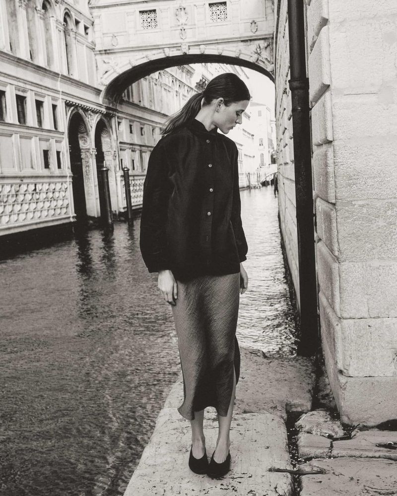 Monochrome portrait of a woman in black jacket and skirt standing on a stone walkway with a canal and buildings in the background.