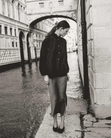 Monochrome portrait of a woman in black jacket and skirt standing on a stone walkway with a canal and buildings in the background.