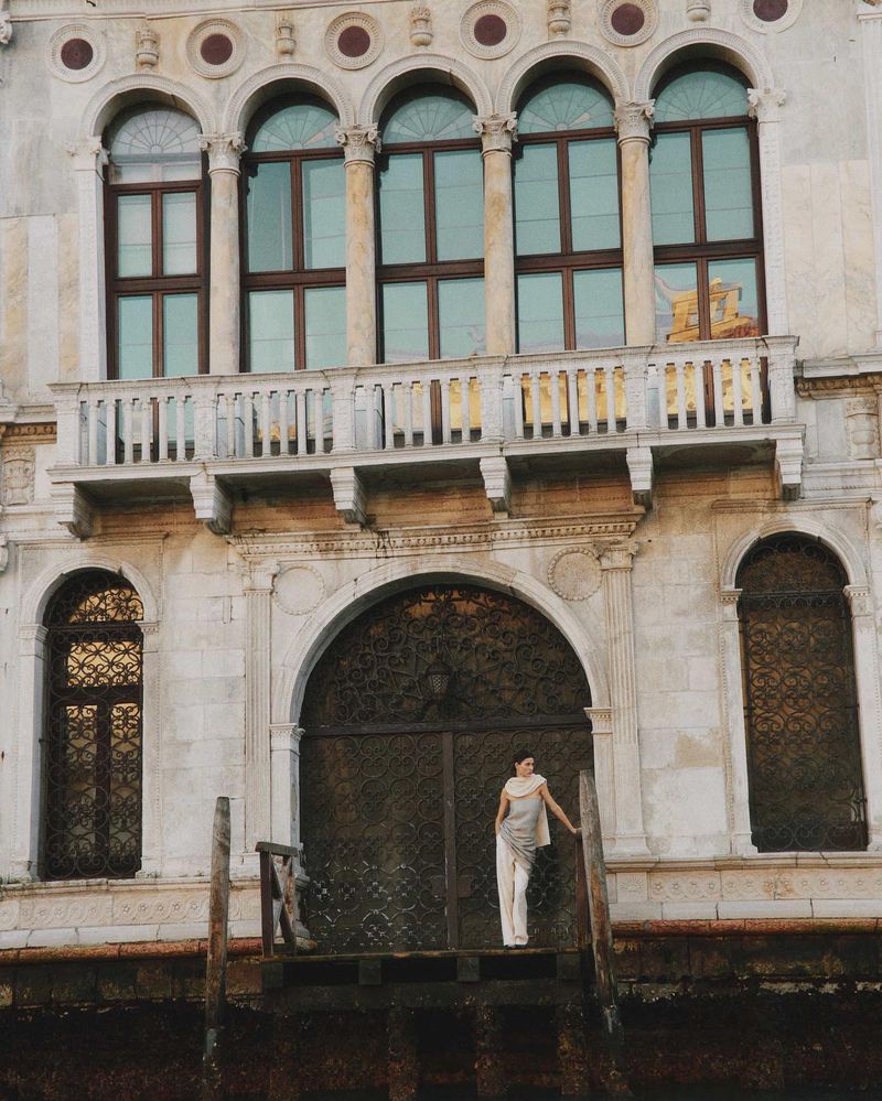 Woman in neutral clothing posing on dock in Venice, Italy, with ornate marble architecture featuring arched windows, columns, and decorative gates.