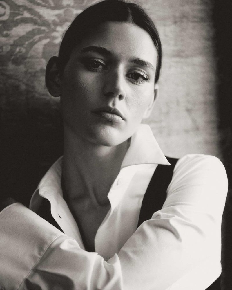 Monochrome portrait of a serious woman with dark hair in a white button-down shirt, looking directly at the camera with arms crossed.