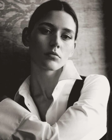 Monochrome portrait of a serious woman with dark hair in a white button-down shirt, looking directly at the camera with arms crossed.