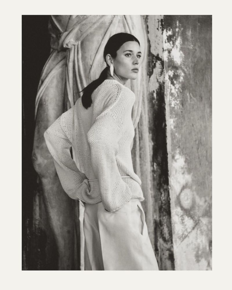 Monochrome portrait of a woman in light-colored knitwear and trousers, posed in front of a worn stone column and sculpture.