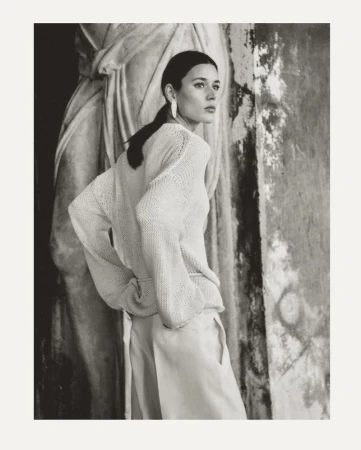 Monochrome portrait of a woman in light-colored knitwear and trousers, posed in front of a worn stone column and sculpture.