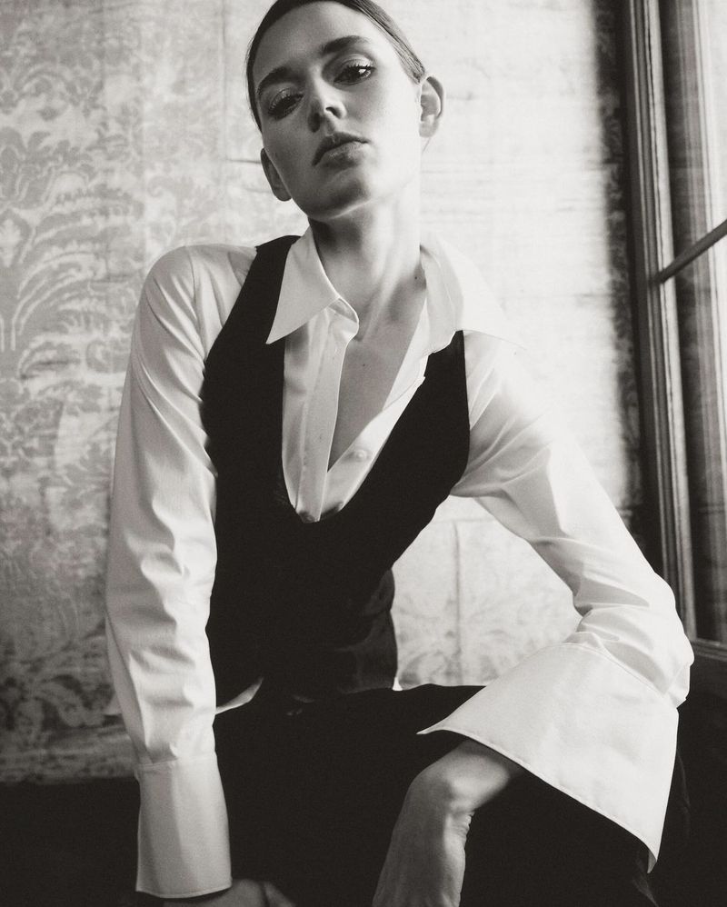Monochrome portrait of a young adult woman in a vest and collared shirt, seated indoors with patterned wallpaper.