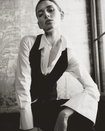Monochrome portrait of a young adult woman in a vest and collared shirt, seated indoors with patterned wallpaper.