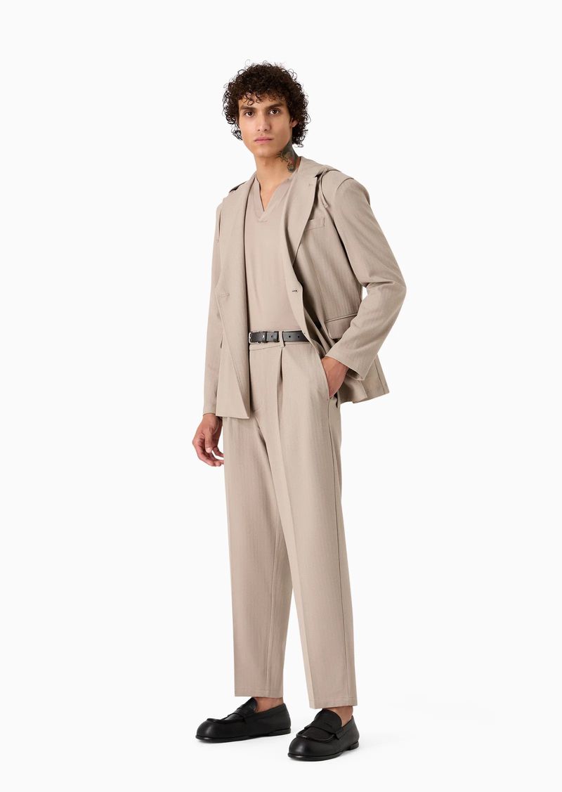 A male model poses in a neutral-tone suit with a V-neck shirt and black loafers against a white background in a full-length fashion shot.