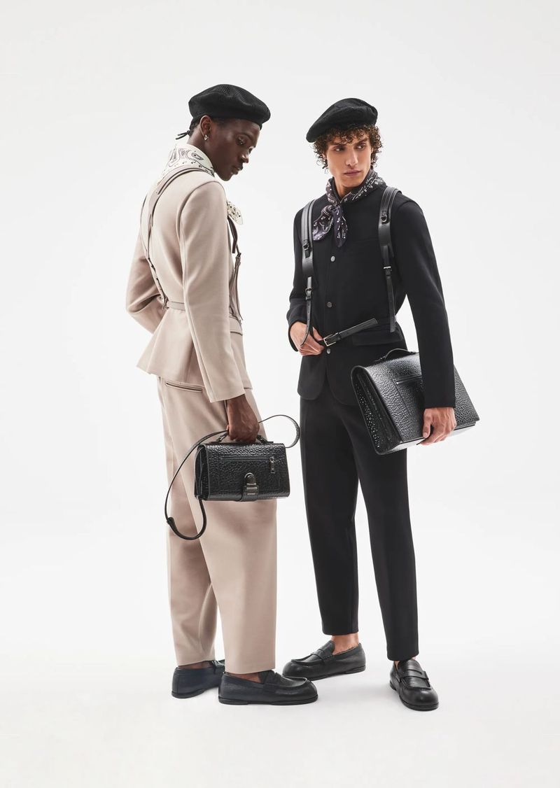 Two male models in berets and tailored suits carrying black leather handbags against a seamless white background.